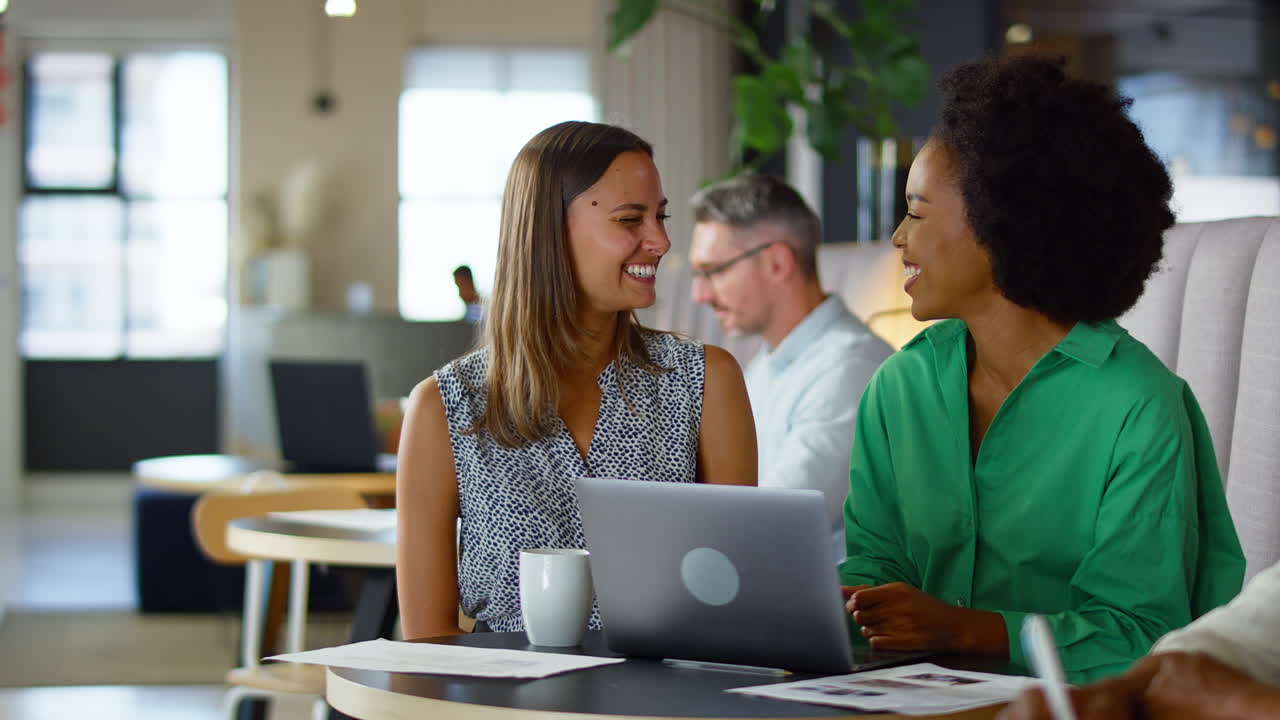 Two Businesswomen Working On Laptop In Informal Seating Area Of Modern Office On Coffee Break