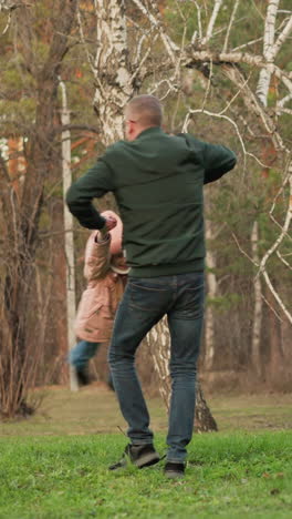 un hombre con una chaqueta verde y pantalones vaqueros azules gira alegremente a una niña en una chaqueta rosa en un campo verde en un parque. alegría pura y energía de un momento lúdico entre un padre y una hija