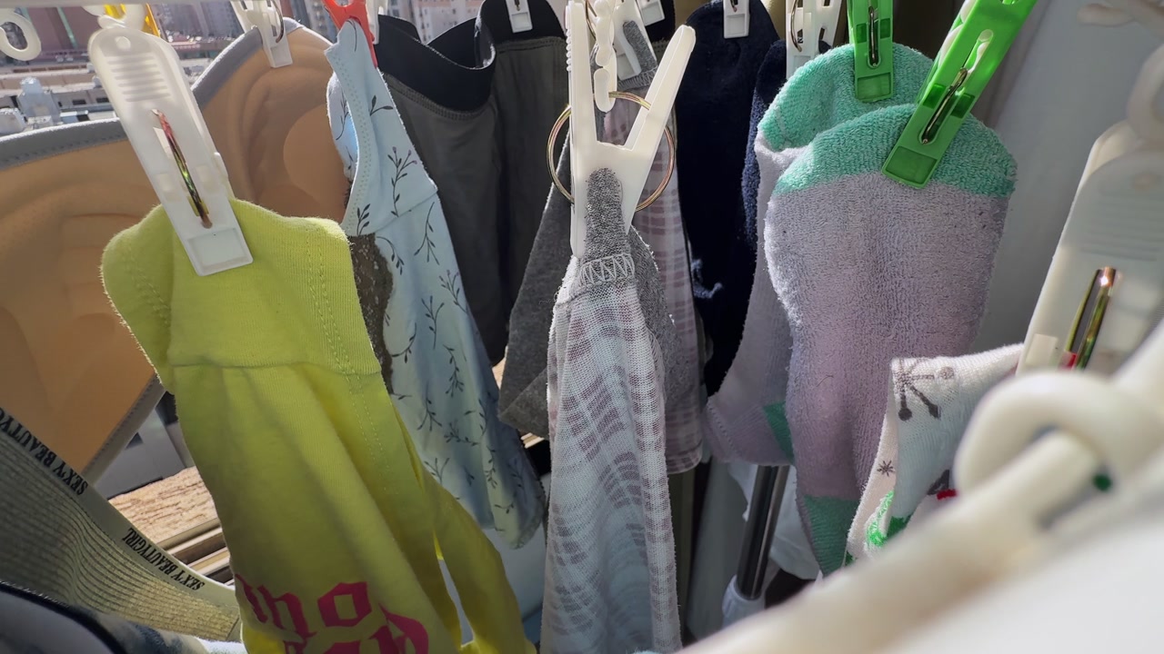 Close-up of socks and small garments clipped to a hanging drying rack by a sunny apartment window, with warm daylight, household laundry details, and an urban residential backdrop.