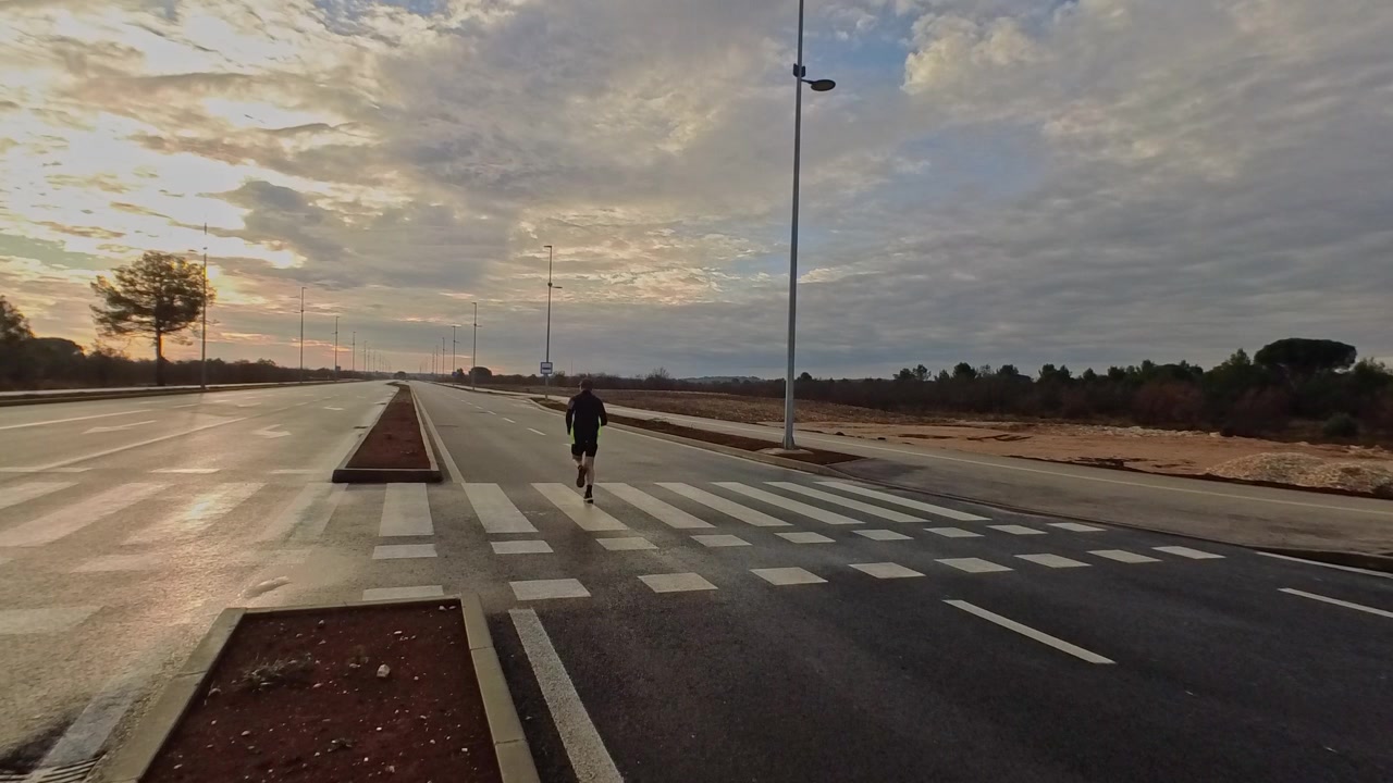Wide shot of an athlete jogging across a crosswalk on a vast, empty road during a cloudy sunrise. Concept of endurance, personal journey, and outdoor fitness.