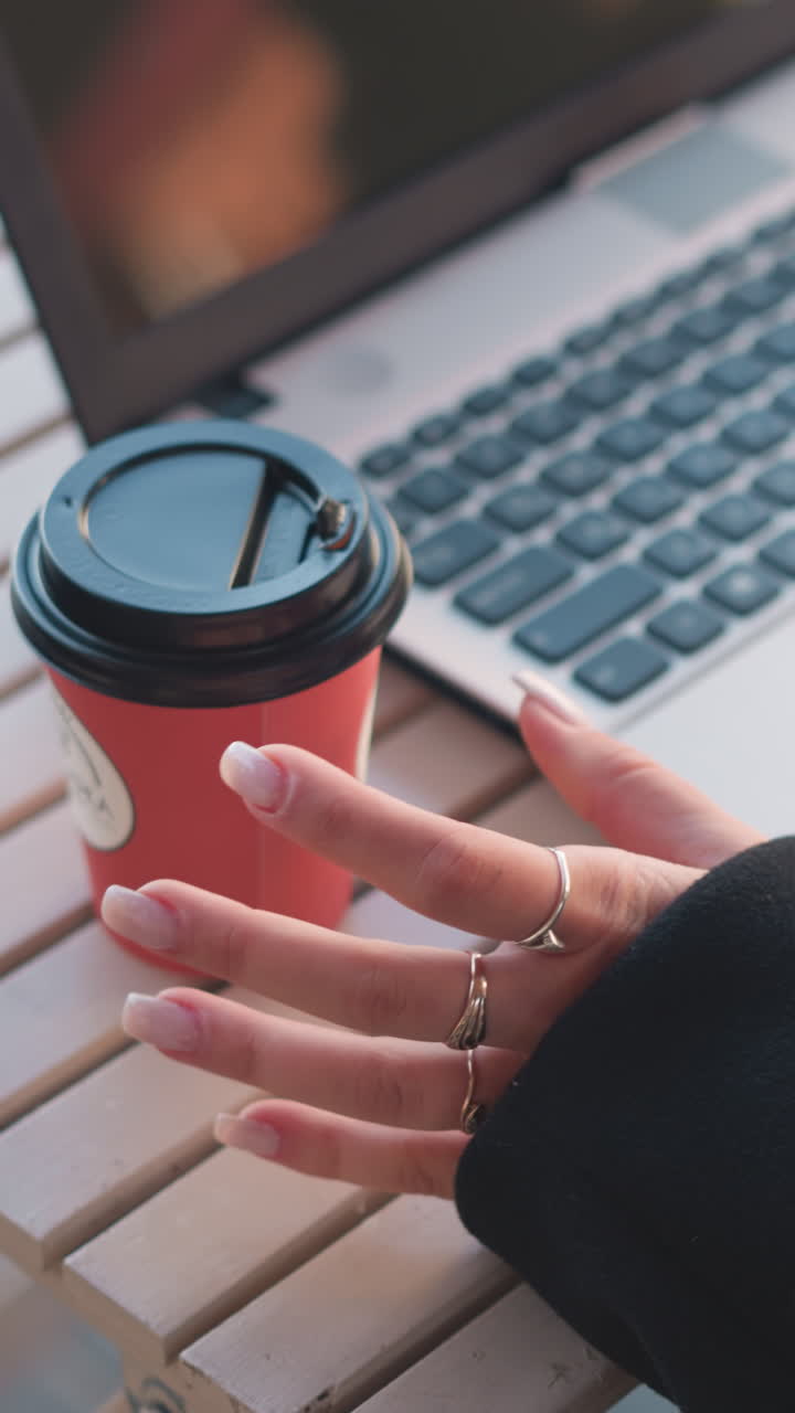 primer plano de una computadora portátil con teclado visible en la mesa. persona con abrigo negro coloca la taza de café en la mesa y comienza a escribir, dedos adornados con anillos, asientos parcialmente visibles en un fondo borroso