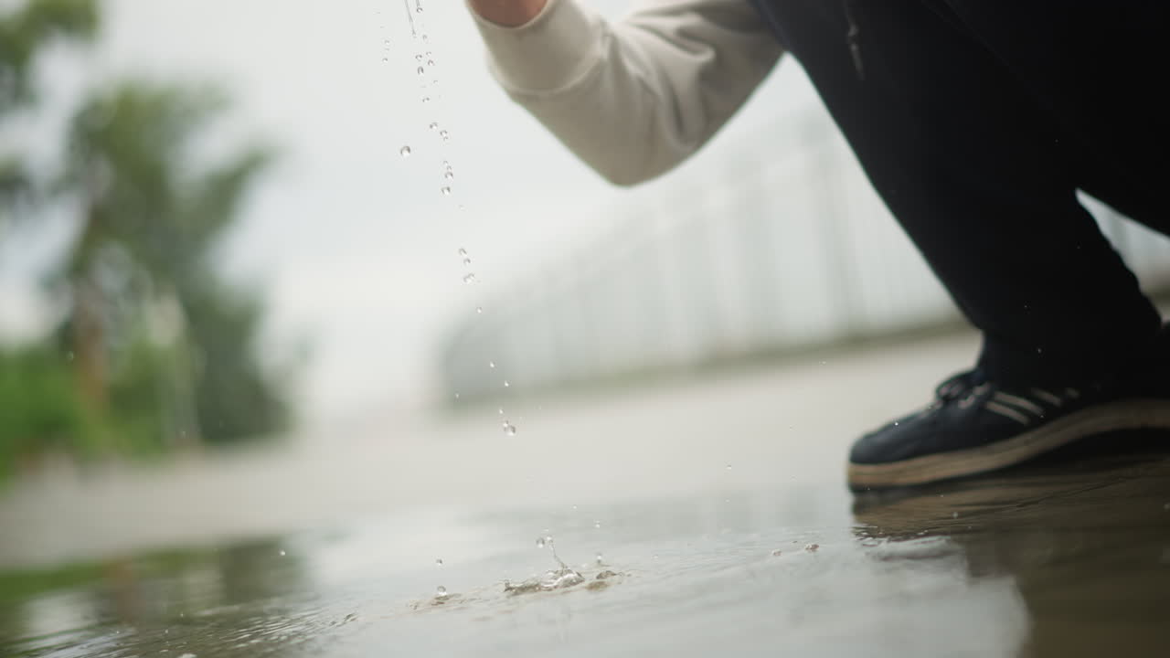 비 오는 길거리의 웅덩이에 손이 닿고, 도시의 통근자가 잠시 멈춰 서 있을 때 어른이 웅크리고 앉아 있는 모습, 부드러운 물보라와 물방울, 스웨터 소매와 운동화가 보이고, 흐린 부드러운 빛, 질감에 얕은 초점