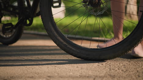 primer plano de las piernas de alguien de pie al lado de un neumático de bicicleta, moviendo suavemente la rueda para comprobar su alineación, la escena se desarrolla al aire libre con un fondo borroso de vegetación y árboles