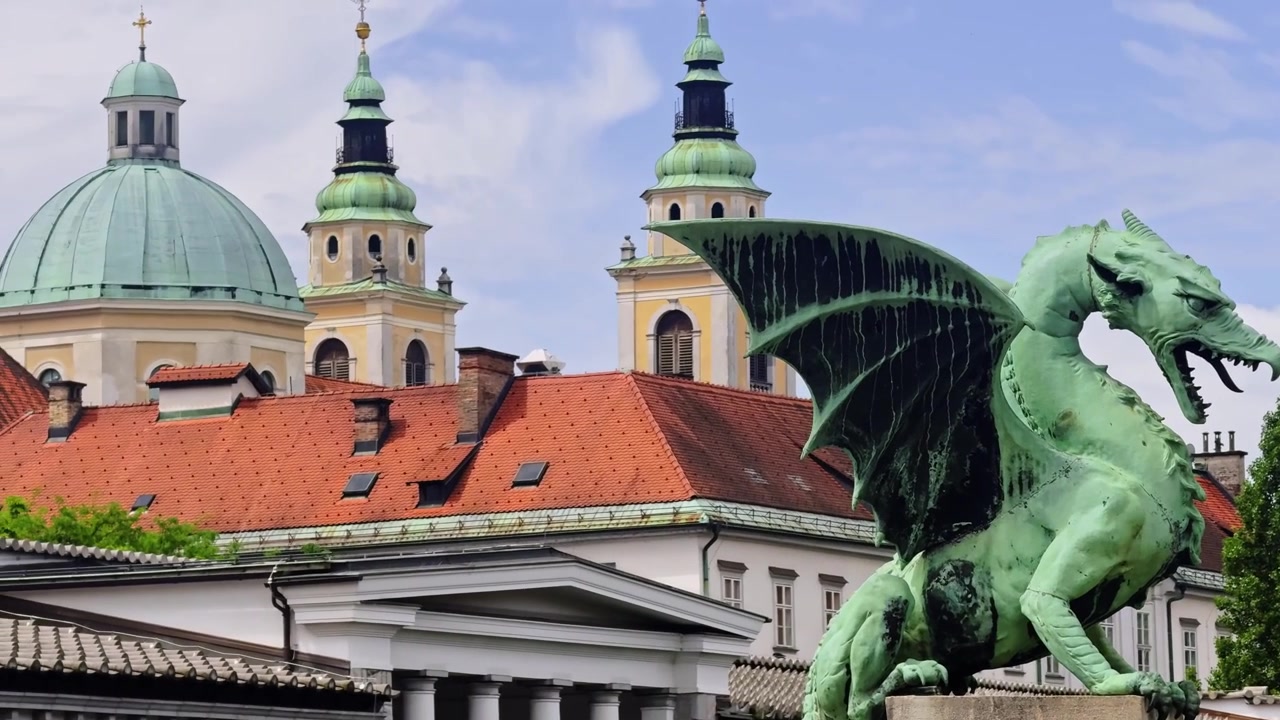 Drageskulptur på Ljubljanas Dragebro, med Ljubljana Domkirke, Sankt Nikolaj Katedralens kupler, røde hustage i den gamle bydel og Plečniks Centralmarked