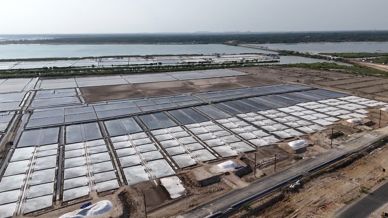 An aerial drone shot of a salt pan in a sun-drenched landscape. The symmetrical ponds and brilliant white salt heaps create a striking visual of industrial-scale nature