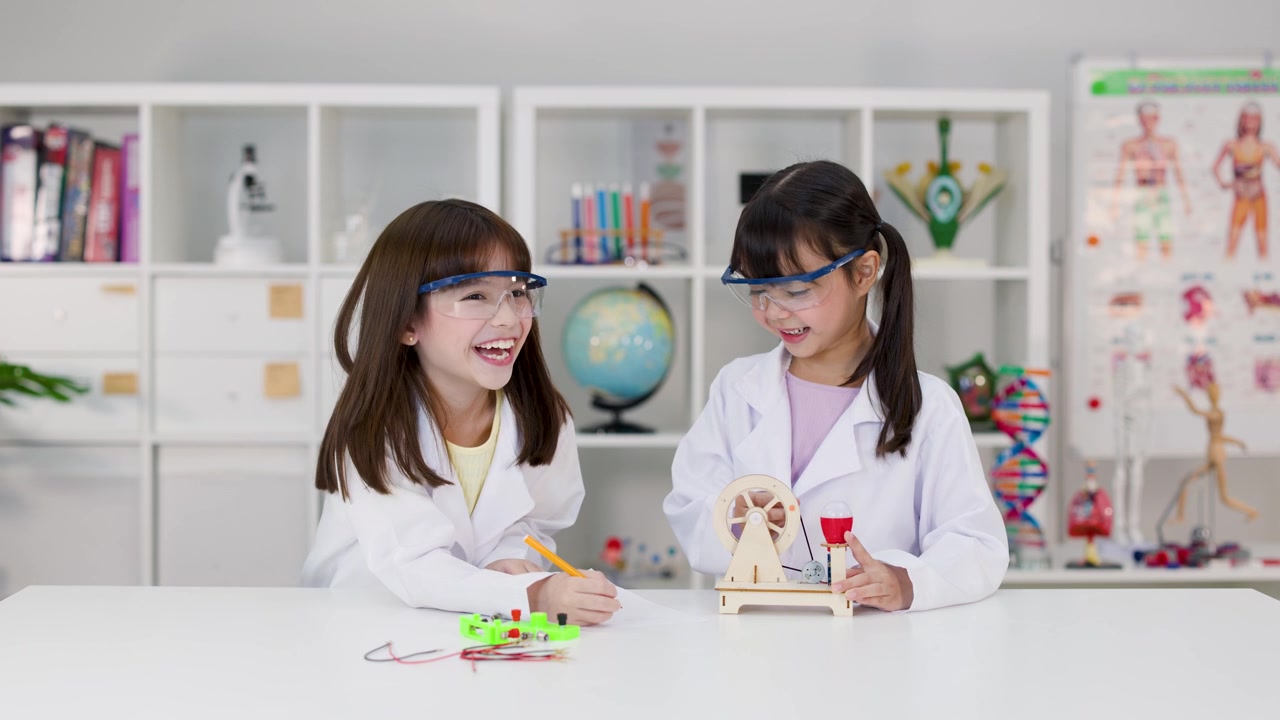 Dua gadis mengenakan jas lab dan kacamata pelindung mengerjakan proyek sains di ruang kelas.