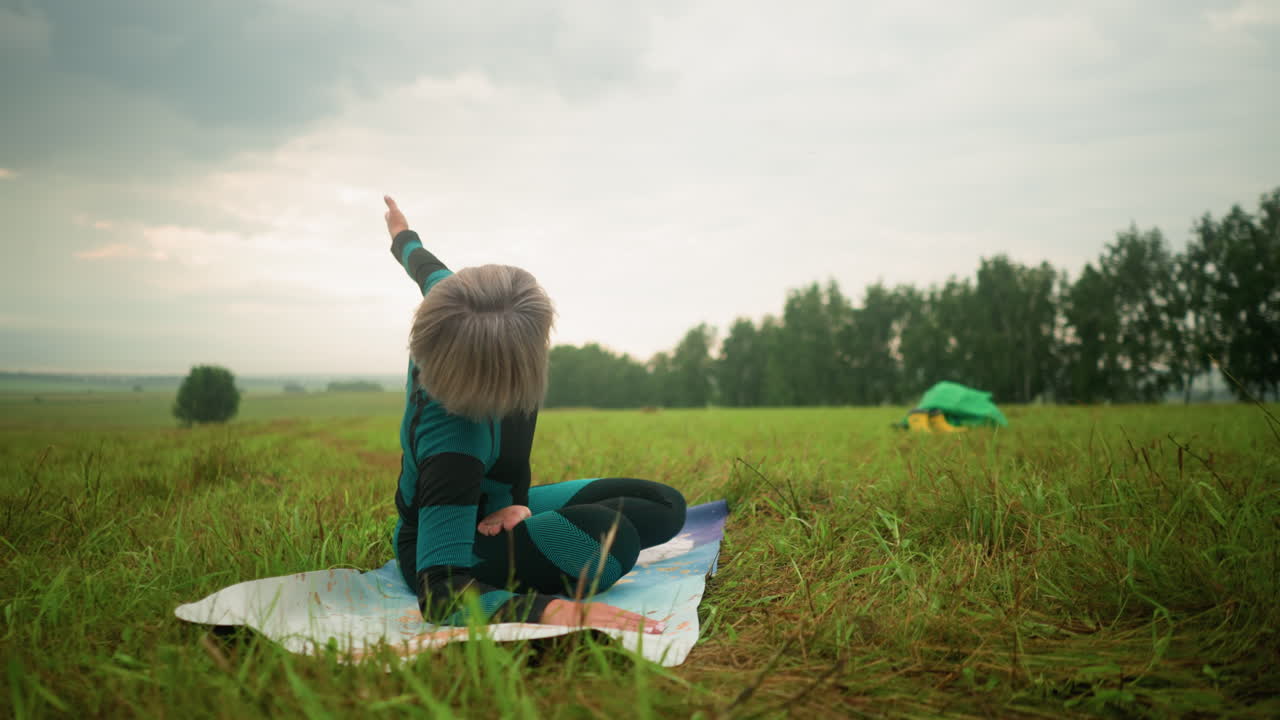 요가 매트 위에 누워있는 여자가 구름이 은 하늘 아래 광활한 풀에서 앉아있는 트위스트 포즈를 연습하고 있으며, 노란색 부츠와 녹색 천이 배경에 바닥에 보이며, 나무가 지평선을 고 있습니다.