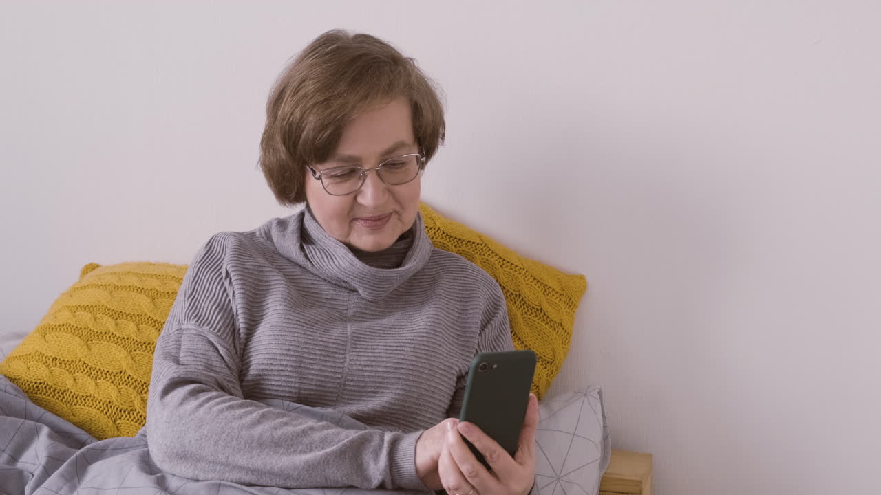 Senior woman with eyeglasses sitting on bed using smartphone