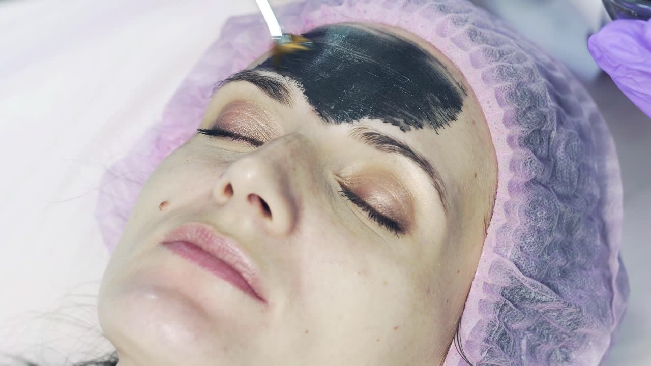 Cosmetologist smears cosmetic mask on the face of the woman in the spa salon. Beautiful female in medical cap receiving black mask on face. Close-up