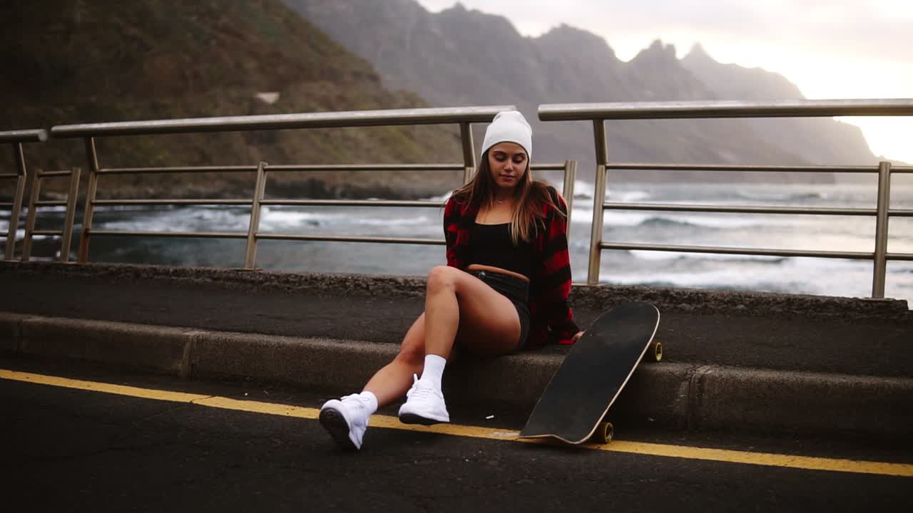 Girl sitting with longboard at the border at coastline asphalt road on background hills and foggy ocean. Relaxing break. Cloudy weather