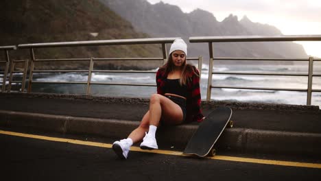 chica sentada con longboard en la frontera en la carretera de asfalto de la costa en las colinas de fondo y el océano brumoso. descanso relajante. clima nublado