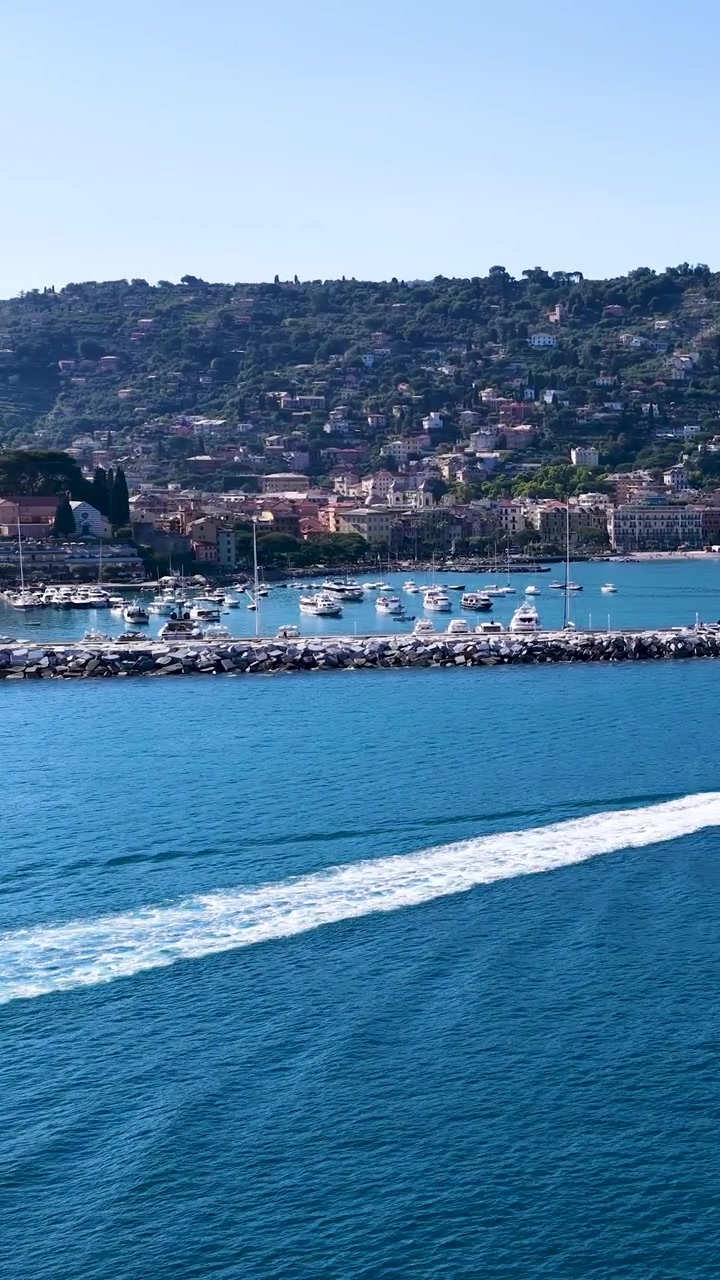 La scia di una barca bianca taglia l'acqua blu con una città costiera e colline sullo sfondo.
