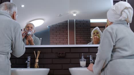 senior grandmother applying facial mask and grandfather shaving with manual razor blade at bathroom