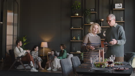 happy grandparents preparing dining table, while in the background their family sitting in living room and talking together 1