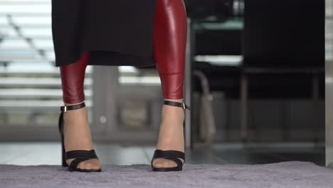 a young woman with sporty sexy legs wears shiny red leggings and high-heeled black stiletto sandals walks into the picture while doing a fashion show, the camera follows her movement