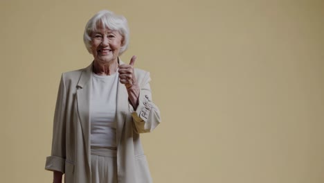 optimistic senior woman signaling thumbs up gesture, white hair and casual suit, expressing confidence and positive emotion against yellow studio background