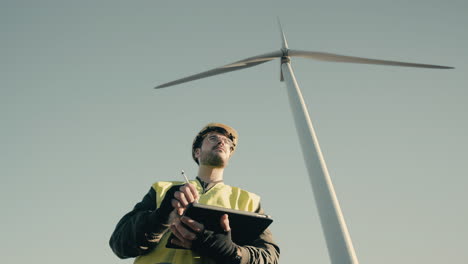 working to promote eco-friendly energy, an engineer in a white helmet and reflective vest uses technology to audit wind turbines in a field of renewable energy generators on a sunny day