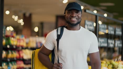 Retrato-De-Un-Trabajador-De-Reparto-Masculino-Feliz,-De-Piel-Negra,-Con-Un-Sombrero-Negro-Y-Una-Camiseta-Blanca,-Que-Lleva-Una-Gran-Bolsa-Amarilla-Sobre-Sus-Hombros-Mientras-Busca-Los-Productos-Necesarios-En-Un-Supermercado.