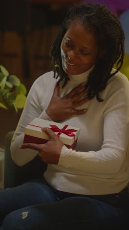 Vertical-Video-Of-Woman-Giving-Female-Friend-Birthday-Present-At-Home-With-Party-Balloons-In-Background