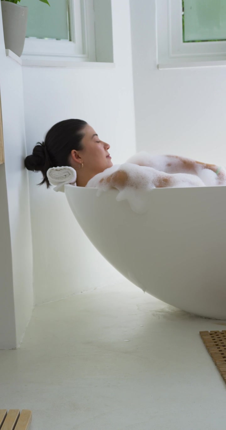 Vertical video: Reclining adult woman relaxing with head on rolled towel in freestanding tub, foam
