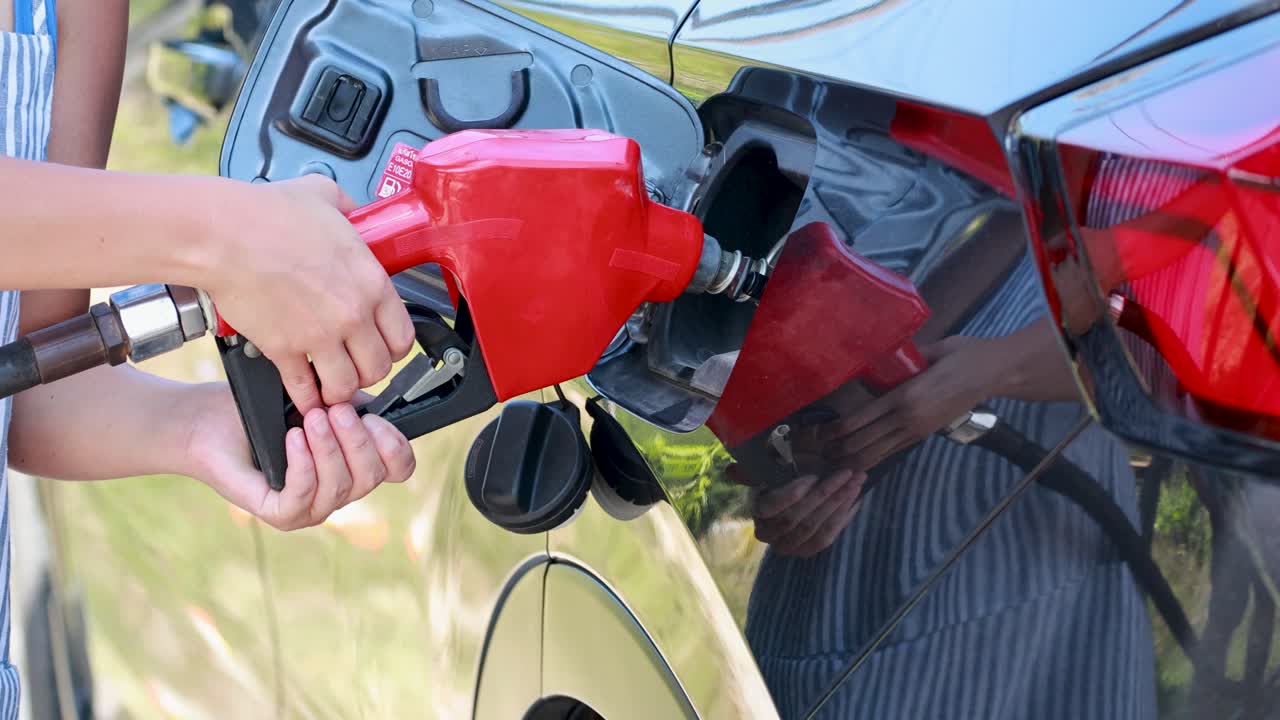 A person refuels a car at a gas station in Phuket, Thailand, under bright daylight, focusing on the fuel nozzle and vehicle