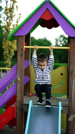 caucasian kid waves on the horizontal bar and goes down by the slide. fun time for a baby at the playground. vertical video.