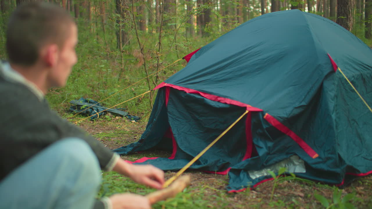 Back view of man holding wooden stick while crouching and hammering peg into ground near pitched tent in forest, surrounded by trees and grass during peaceful daytime outdoor camping setup