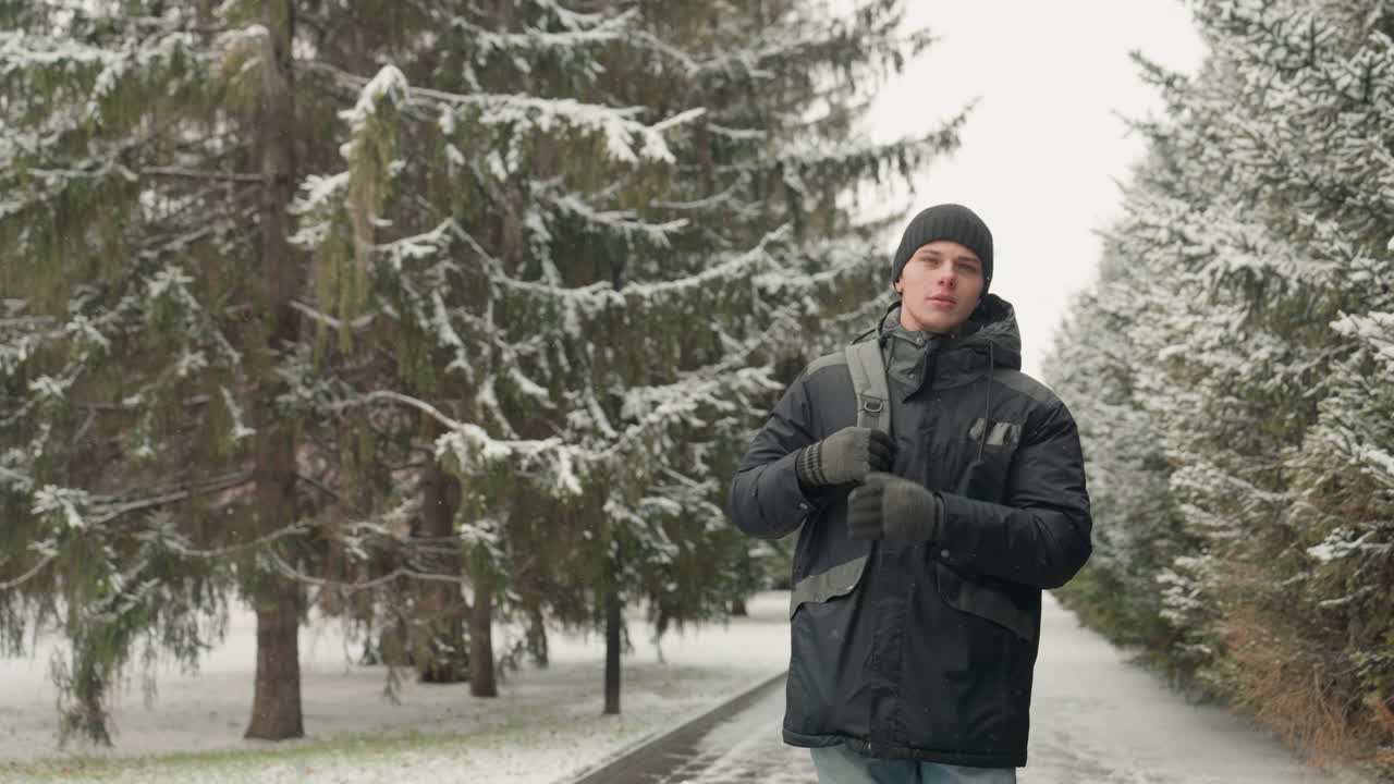 Man adjusting backpack on snowy path, tightening gloves and pulling beanie while walking past frosted pine trees, breath visible in cold air, wearing black parka and jeans, urban park walkway