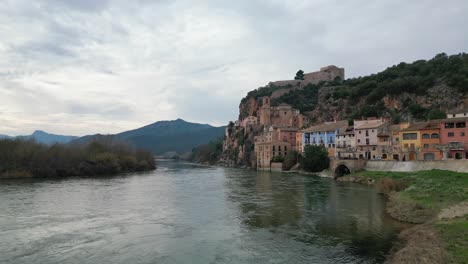 Río-Que-Fluye-Por-La-Histórica-Ciudad-De-Miravet-En-Tarragona,-España,-Con-Montañas-Al-Fondo,-Cielo-Nublado