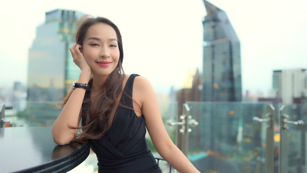 With the Bangkok skyline behind her, a light breeze blows through the hair of an attractive young woman in a little black dress sitting at a rooftop bar