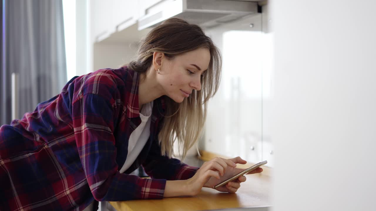 Blonde woman using mobile phone in the kitchen leans on counter