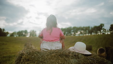 back view of young girl in pink sitting on hay bale while boy wearing glasses focuses on hay, sun hat rests beside her as they enjoy peaceful countryside with open field and trees under cloudy sky