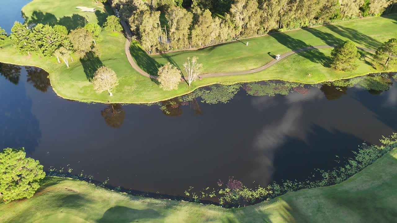 Drone footage captures a serene golf course with lush greenery and a reflective lake under bright sunlight