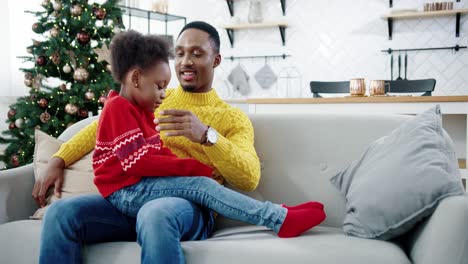 Portrait-Of-Father-Speaking-With-Little-Cute-Child-While-Sitting-On-Sofa-In-Christmassy-Decorated-House