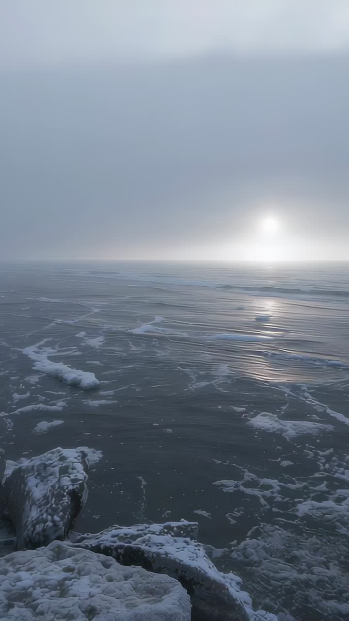Vertical video: Shifting fog, sun and sea casting dim light on snow-dusted boulders at rocky shore