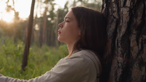 woman rests near tree trunk in woods at sunset. young lady feels emptiness with anxiety thinking about ways to improve mental health. finding sense of life