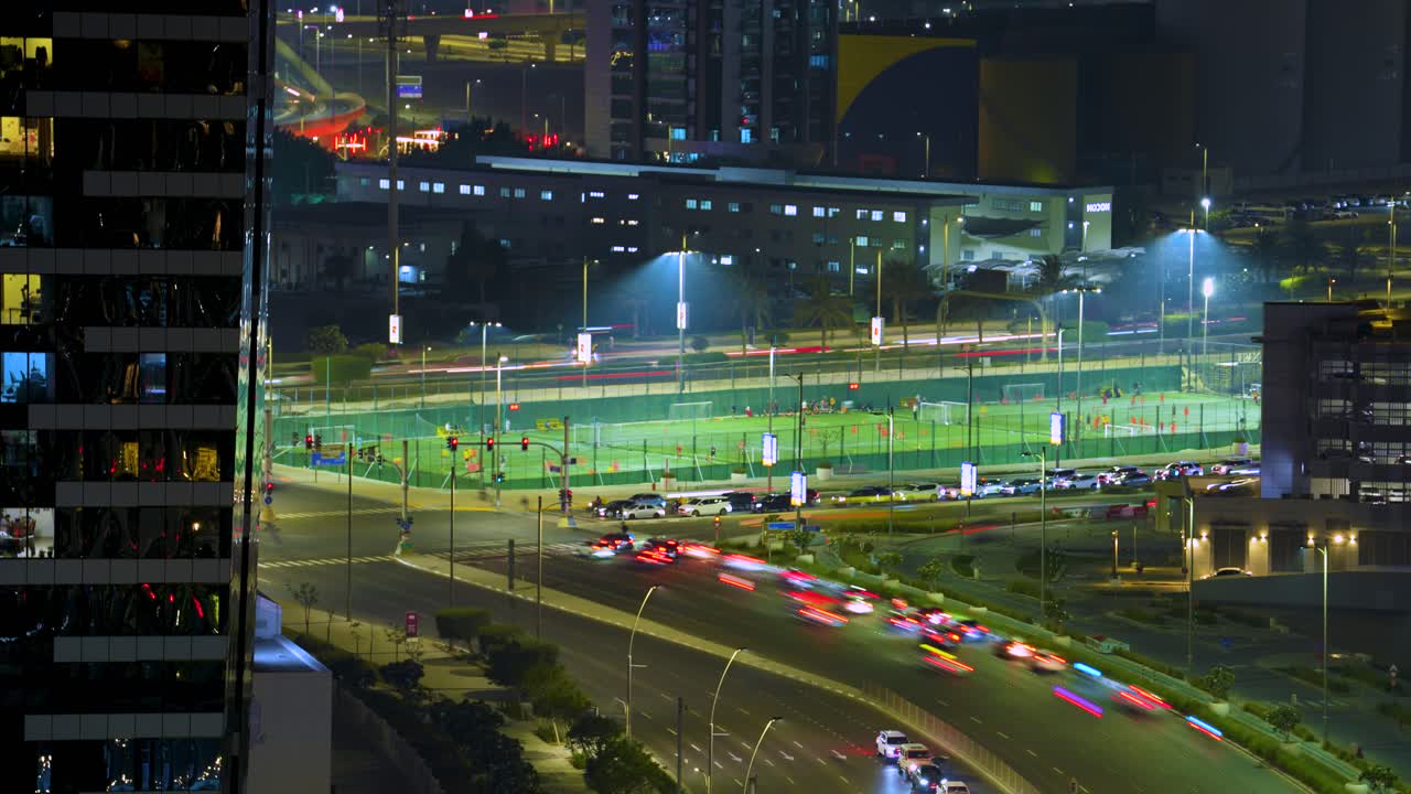 Abu Dhabi night traffic intersection with vibrant city lights in a time lapse. A roadside football or soccer pitch hosts an amateur scrimmage, showcasing urban sports life in the United Arab Emirates