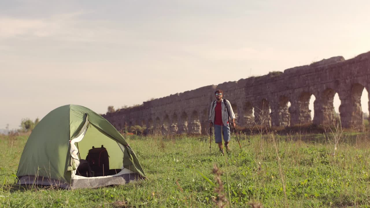 캠핑 텐트 밖에서 배을 고 있는 젊은 남자가 잔디 위에 지이를 들고 맨발로 걸어가며 로마 아쿠에듀크 앞에 서 있는 파르코 델리 아쿠에두티 공원 유적에 서서히 서서히 고 있다.