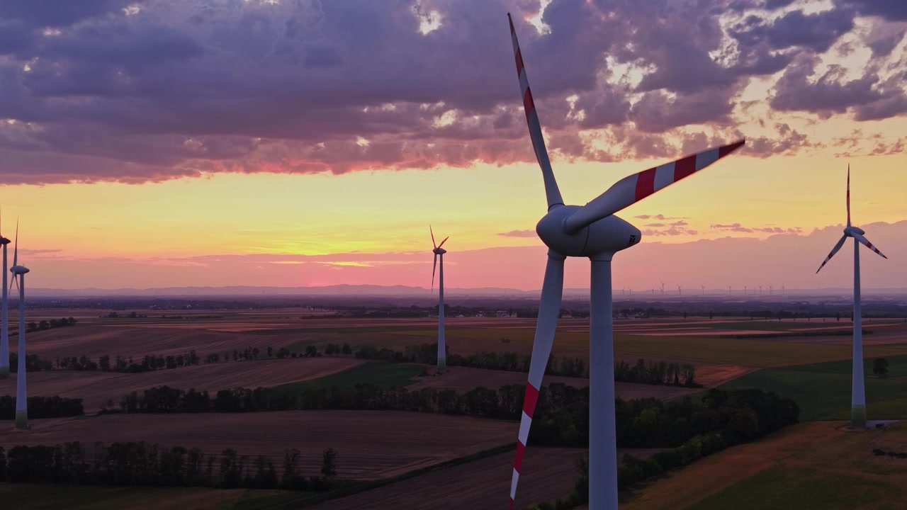Wind turbines stand in fields during sunset with clouds in the sky