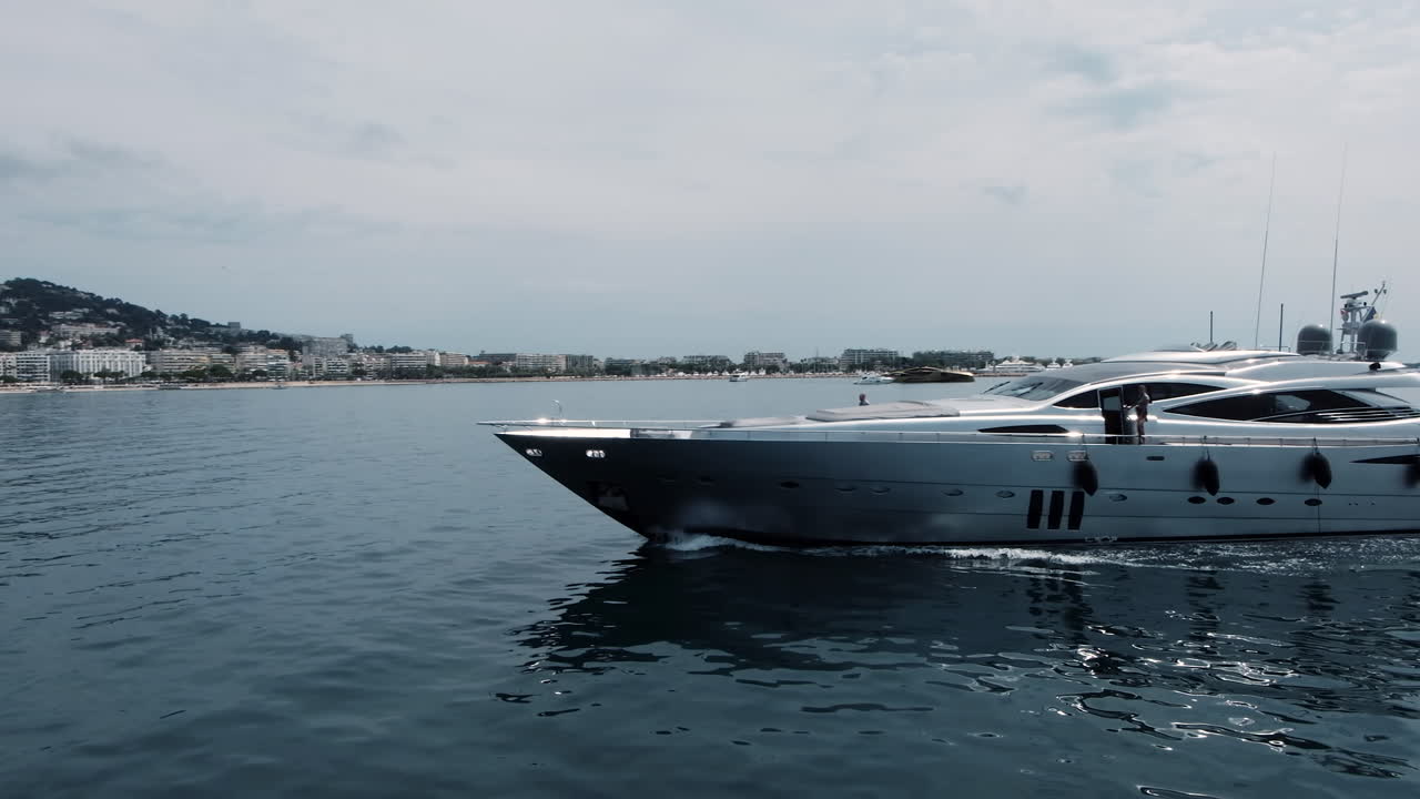Elegant black and white yacht glides along the calm sea near the French Riviera coast, with modern buildings and sailboats in the background