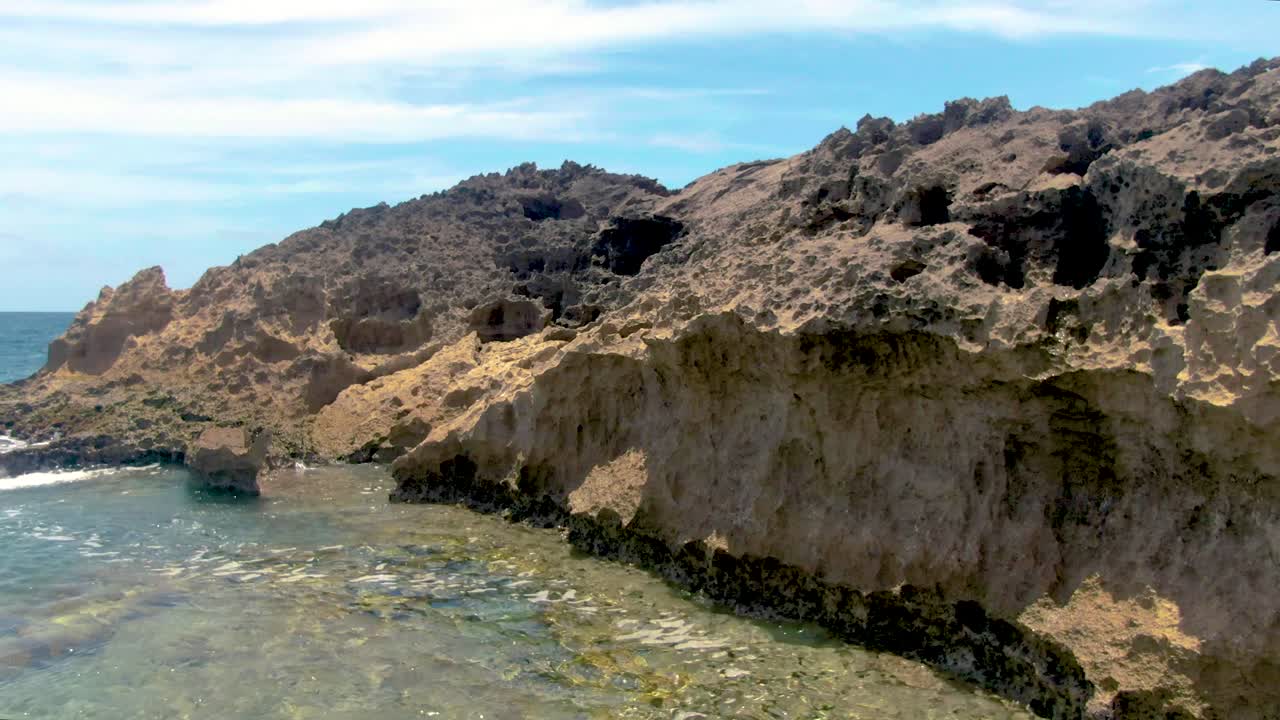 A drone POV flying above turquoise seawater through a narrow channel of jagged rocks to reveal the ocean