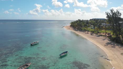 4k-Aéreo-Que-Establece-Una-Toma-De-Agua-Azul-Clara-A-Lo-Largo-De-La-Playa-Con-Botes-De-Remos-Y-Palmeras-En-Pasamontañas,-Mauricio