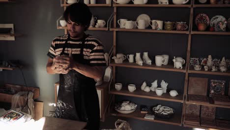 young male potter kneads clay against background of a rack with ceramic blanks