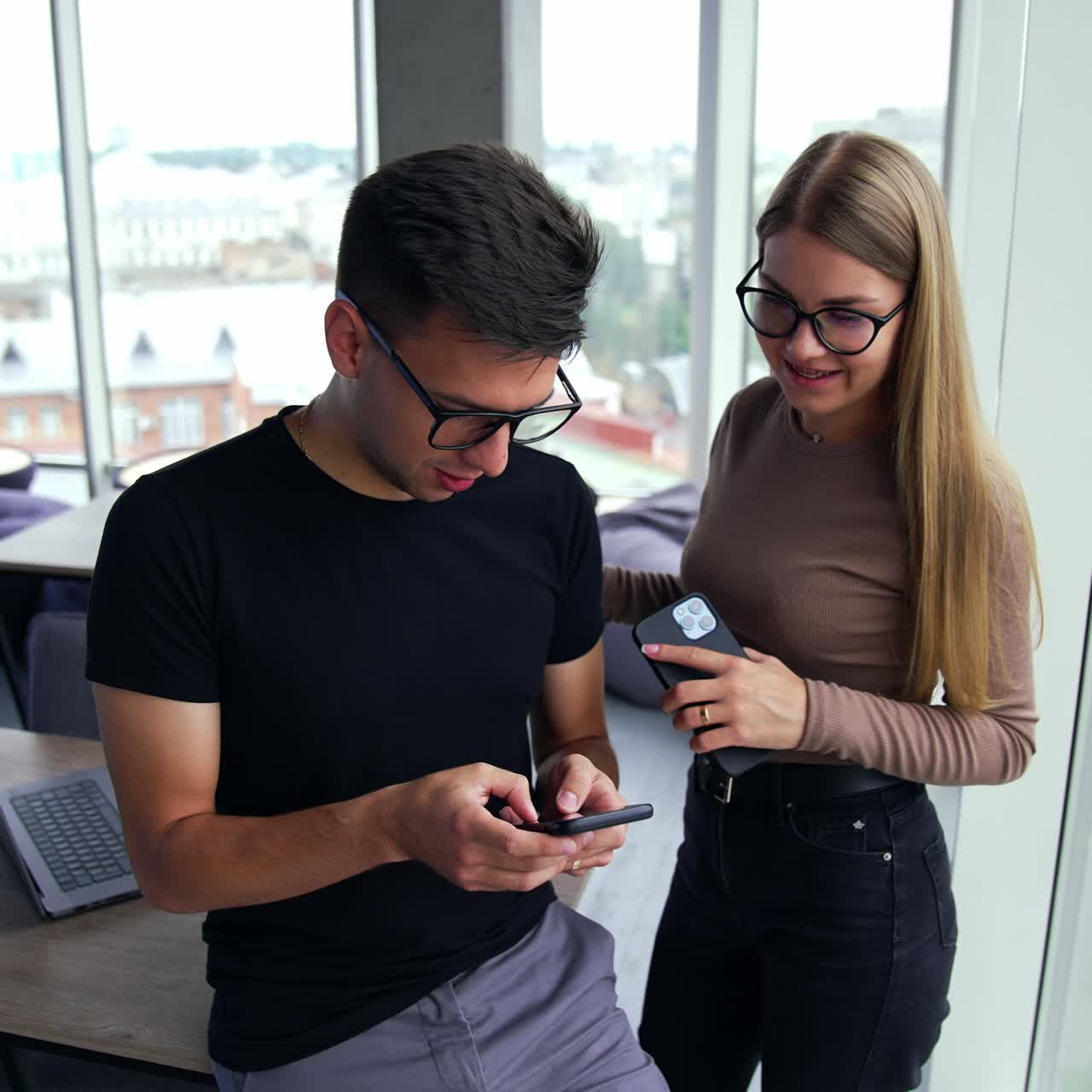 Diverse colleagues standing near window in the office. Male and female using phones. Cityscape in window at backdrop in blur