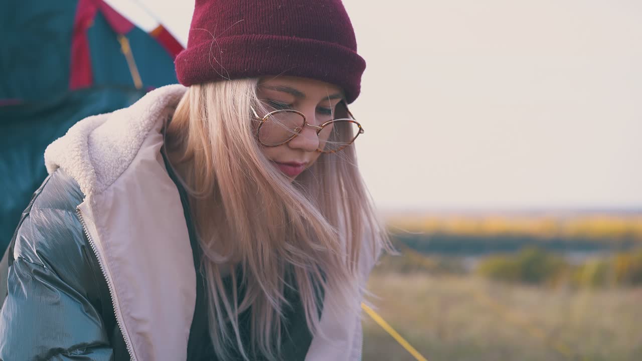 una bella ragazza con occhiali e cappello si siede vicino alla tenda blu al tramonto