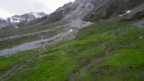 Luftaufnahmen-Von-Drohnen,-Die-über-Eine-Almwiese-Fliegen,-Bevor-Sie-Sich-Winden-Und-Durch-Eine-Schroffe,-Von-Gletschern-Geformte-Berglandschaft-Mit-Schneeflecken-Und-Einem-Wanderweg-In-Der-Schweiz-Aufsteigen