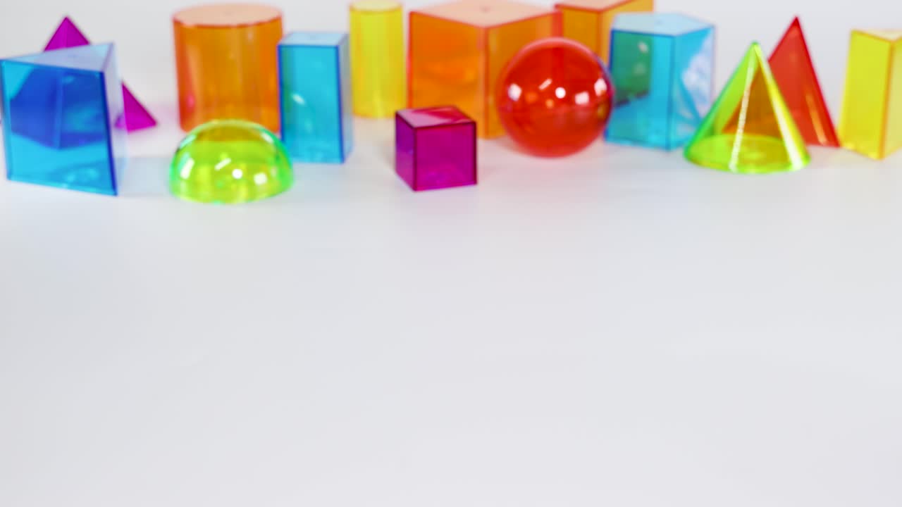 A hand rolls a red sphere among colorful geometric shapes on a white surface, showcasing playful interaction and vibrant colors