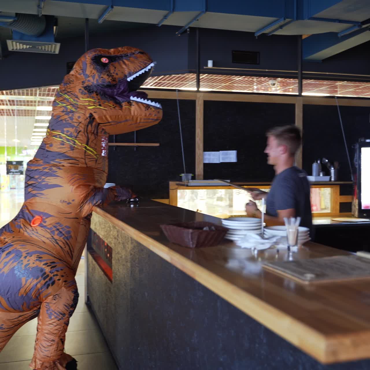 Person in dinosaur inflatable costume visiting the café. Orange dinosaur comes up to a bar stand and is given a menu by the bartender