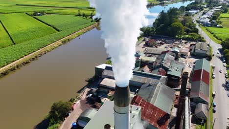 drone footage captures steam rising from a factory chimney surrounded by lush fields and a river under clear skies