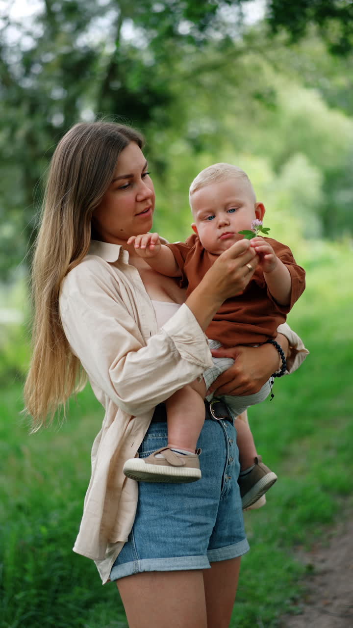 Blond Caucasian woman with long hair holds a toddler boy in hands. Blond kid giver a clover flower to his mom to smell. Vertical video.